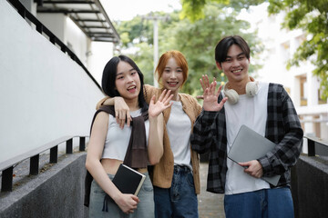 Three young students happily walking on campus, waving and chatting with laptops and books, exuding togetherness and friendship in a modern urban setting on a sunny day