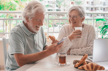 Smiling elderly couple enjoying a morning breakfast together at a cozy outdoor balcony with croissant and coffee and tea, authentic serene retirement lifestyle