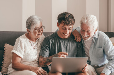 Modern senior couple learning computer technology from experienced young grandson, handsome smiling boy teaching to his grandparents how to use computer, make online shopping, book travel and vacation