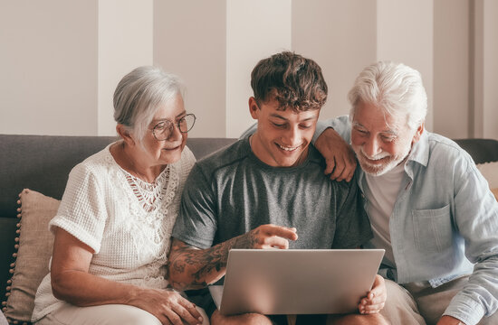 Modern senior couple learning computer technology from experienced young grandson, handsome smiling boy teaching to his grandparents how to use computer, make online shopping, book travel and vacation