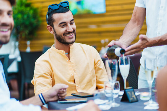 Young diverse friends enjoying dinner party at restaurant, waiter serving wine, multicultural gathering, social event, casual dining experience