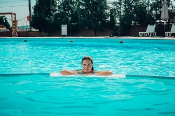 A woman is swimming in a pool. She is wearing a green top and is smiling. The pool is blue and has a few chairs around it.