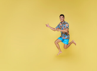 Full body young asian tourist man wear beach shirt jumping and pointing finger isolated on yellow copy space background studio. Summer vacation sea rest sun tan concept