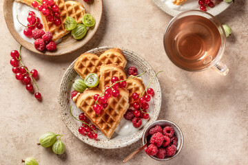 Homemade waffles with summer berries and yogurt served for sweet breakfast with tea on beige background. Heart shaped waffles top view