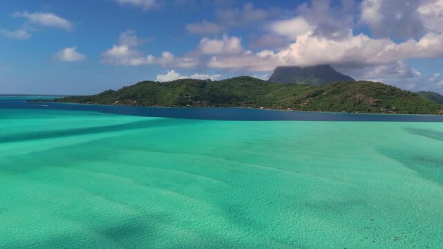 Beautiful fluorescent blue, turquoise and azure water surrounds the tropical island of Bora Bora in French Polynesia, The low flying drone is moving over the south pacific ocean toward land.
