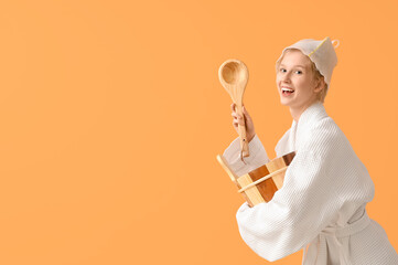 Naklejka premium Happy young woman in white robe and sauna hat holding wooden bucket and ladle for spa relaxation