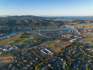 Arial wide of town with river running through along bay, lots of blue water and green trees, golden hour sunset shining