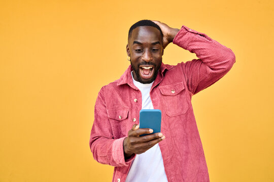 Excited Black man looking at smartphone with wow reaction isolated on yellow. Happy African young gen z guy holding cellphone reading message on cell phone. Amazed user winner using mobile phone