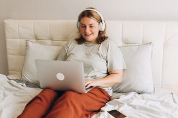 Nice beautiful 30s woman with headphones working at the laptop and sitting on the bed at home. Concepts of education, work and comfort