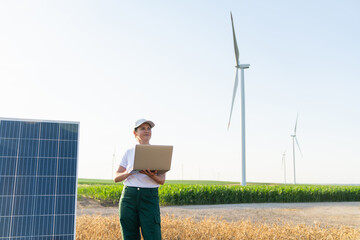 Woman farmer wearing white cap and t-shirt with laptop stands next to solar panel. Wind turbines in the background