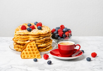Stack of Belgian waffles, berries, honey and red cup of coffee: delicious summer breakfast on marble table. Heart-shaped waffle and mix of raspberries and blueberries as topping.