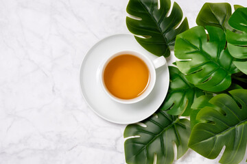 White cup of tea on marble background with green Monstera leaves. Top view, copy space for text