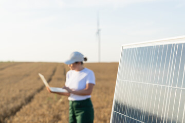 Woman farmer wearing white cap and t-shirt with laptop stands next to solar panel. Wind turbines in...