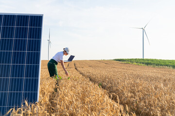 Woman farmer wearing white cap and t-shirt with laptop stands next to solar panel. Wind turbines in...