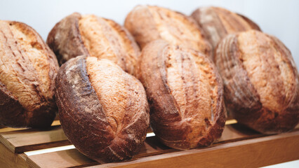A row of bread loaves are on a wooden shelf. The loaves are all different sizes and shapes, but they all have a similar appearance. The bread looks fresh and inviting