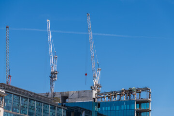 Close-up low angle view of group of working cranes on construction site of modern high-rise apartment or office building against blue sky in sunny day. Copy space. Development theme.