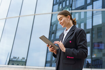 Fototapeta premium A young professional woman, focused on her tablet, stands confidently against a modern glass building backdrop.