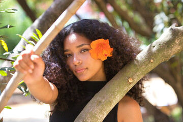 portrait of a beautiful young Latin woman with large, dark eyes and curly hair. The girl wears an orange flower over her ear and clings to the trunk of the tree, the woman looks serene and peaceful.