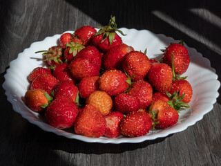 Close-up of detailed, red, ripe, organic strawberries of different sizes with green leaves on a white plate on grey table