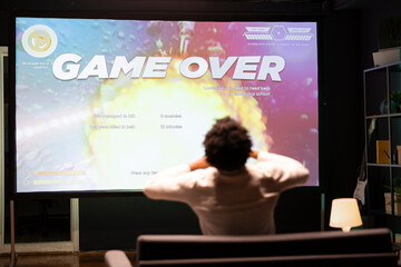 Gamer upset at seeing game over message on ultrawide TV screen playing arcade space shooter videogame. Man spending time at home on gaming system, frustrated after losing online competition