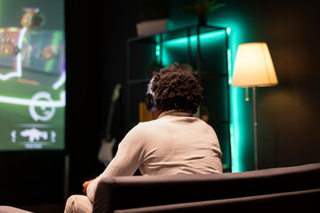 BIPOC man playing SF FPS videogame on widescreen smart TV, having fun in neon lit apartment. Gamer...