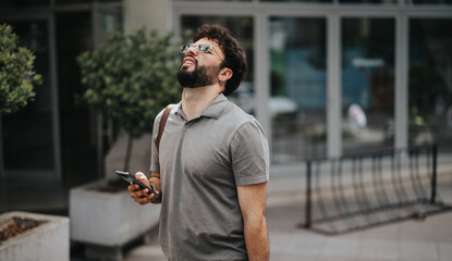 Disappointed businessman with beard holding a smart phone and looking up while standing outside modern building in casual attire.