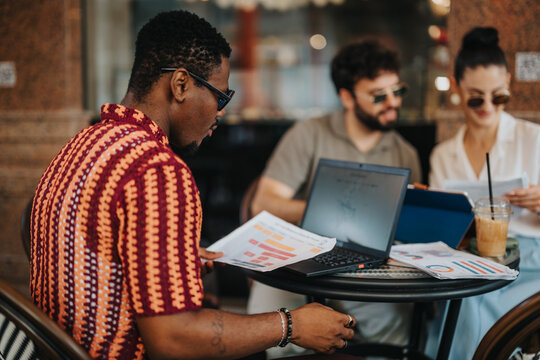 Team of colleagues holding a business meeting in a coffee shop, reviewing documents and charts on laptops and tablets. Collaborative discussion over coffee and printed graphs.