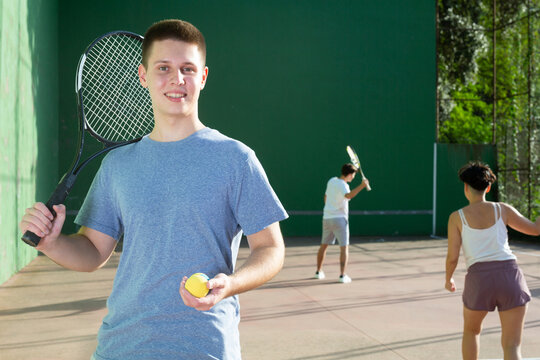 Portrait of positive caucasian boy standing on frontenis court, holding racket and ball