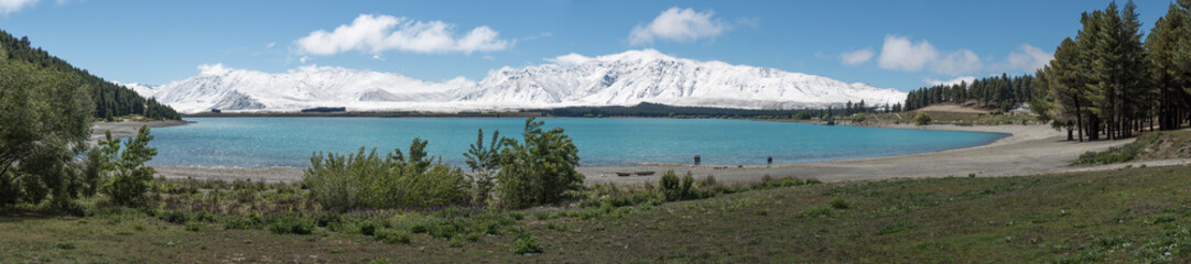 Wide panorama of the turquoise waters of Lake Tekapo, New Zealand. Snow-capped mountains in the background and gravel beach in the foreground.
