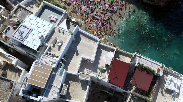 Downward angle drone shot of Lama Monachile beach on a sunny day in Puglia, Italy. Starting on buildings then revealing people enjoying the clear blue waters.