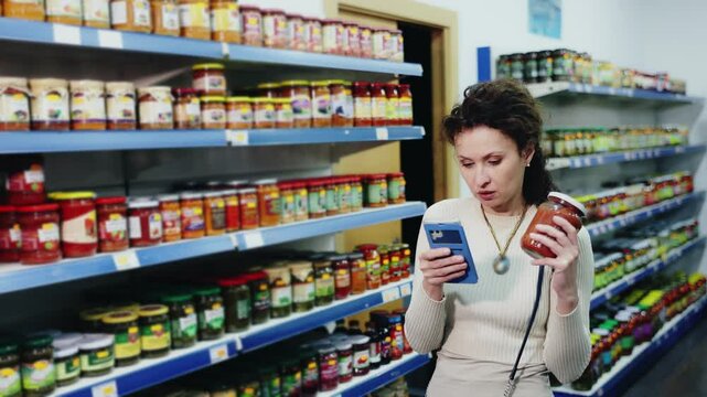 Woan in store of Russian goods photographs jar canned lecho - stewed salad made from bell pepper, tomato sauce. Buyer scan QR code compares item in store and in mobile application 