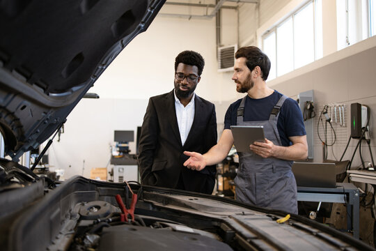 Mechanic explaining car issues to businessman in workshop. Professional technician using tablet to demonstrate vehicle diagnostics. Businessman in suit listening and engaging with mechanic.