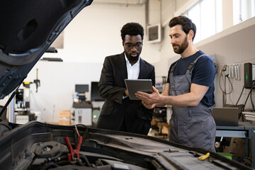 Mechanic wearing overalls discussing car repair details with businessman using digital tablet in...