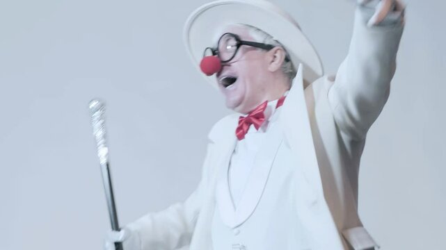 Video of dancing clown. An elderly man in a white coat and hat with walking stick.