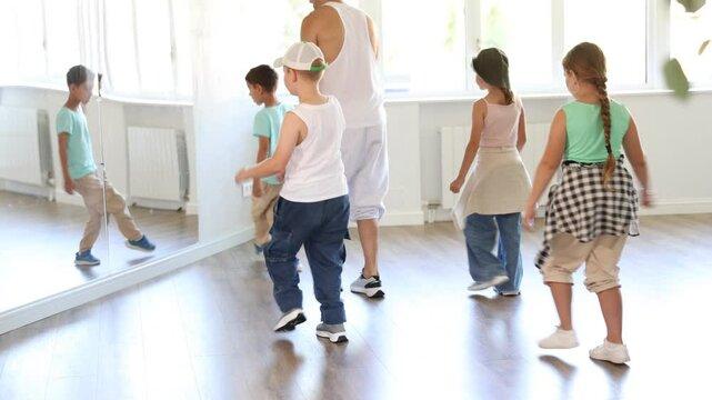 Preteen children enthusiastically following male instructor moves during group hip-hop lesson in sunlit mirrored studio, mastering modern street dance choreography. High quality 4k footage