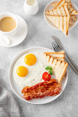 Breakfast with fried eggs, bacon and toasts on a white plate with cup of coffee on a stone background. Top view, selective focus.