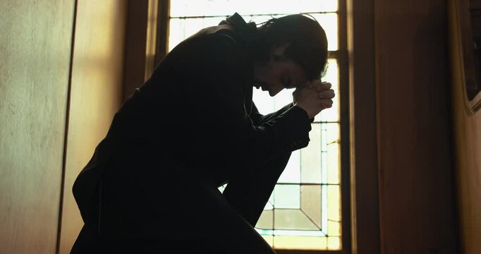 Young man in old church building in front of stained glass window kneeling, praying, worshipping. Christianity, worship, and prayer concept.
