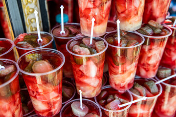 A close-up shot of many plastic cups filled with red and green pickled food. The food is stacked in a market stall, and some of the cups have small white sticks sticking out of them.
