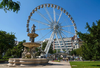 Naklejka premium Budapest, Hungary - September 2, 2023: Low angle and summer view of Danubius Fountain and tourists on the square against a ferris wheel 