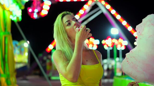 Woman eating a pink cotton candy in an amusement park at night alone