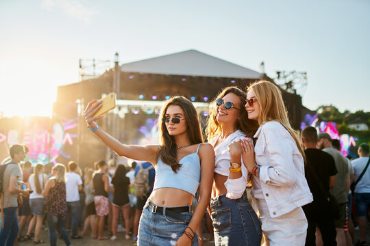 Group of happy young women friends take selfie at sunny beach music fest. Girls in trendy summer outfits enjoy live concert, party atmosphere. Youth culture, friendship, outdoor entertainment moment.