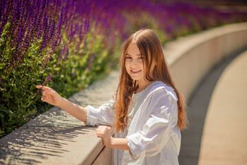 A young girl is basking in the warm rays of the sun, enjoying the beauty of the park, overgrown with bright purple sage. Her white shirt and blue trousers complete the landscape