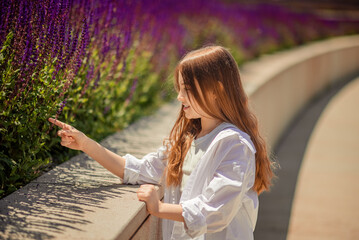 A young girl is basking in the warm rays of the sun, enjoying the beauty of the park, overgrown with bright purple sage. Her white shirt and blue trousers complete the landscape