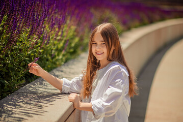 A young girl is basking in the warm rays of the sun, enjoying the beauty of the park, overgrown with bright purple sage. Her white shirt and blue trousers complete the landscape