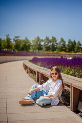 A young girl is basking in the warm rays of the sun, enjoying the beauty of the park, overgrown with bright purple sage. Her white shirt and blue trousers complete the landscape