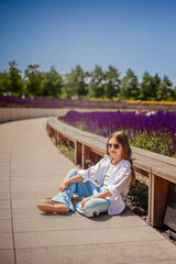 A young girl is basking in the warm rays of the sun, enjoying the beauty of the park, overgrown with bright purple sage. Her white shirt and blue trousers complete the landscape