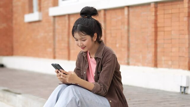 A high school student sits on a pavement against a red brick wall, looking at her phone. She wears a pink shirt, light blue jeans, and a brown jacket. She is absorbed in her phone