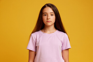 Obraz premium Portrait of beautiful young girl, teen with long brown hair, pink t-shirt, standing with calm expression against yellow studio background. Concept of human emotions, childhood, youth, education