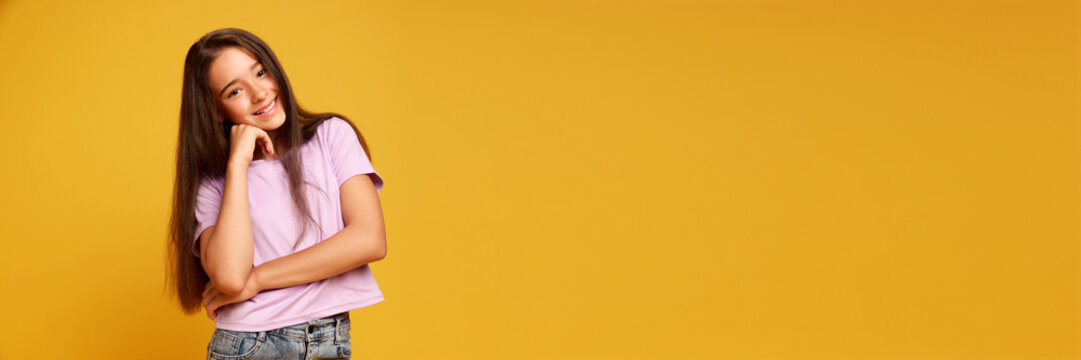Portrait of pretty teen girl with brown long hair, wearing pink t-shirt, standing with cheerful smile against yellow studio background. Concept of human emotions, childhood, youth, education. Banner