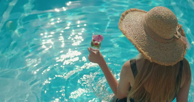  Young woman in a hat holding a cool drink, sitting on the edge of a pool on a hot summer day